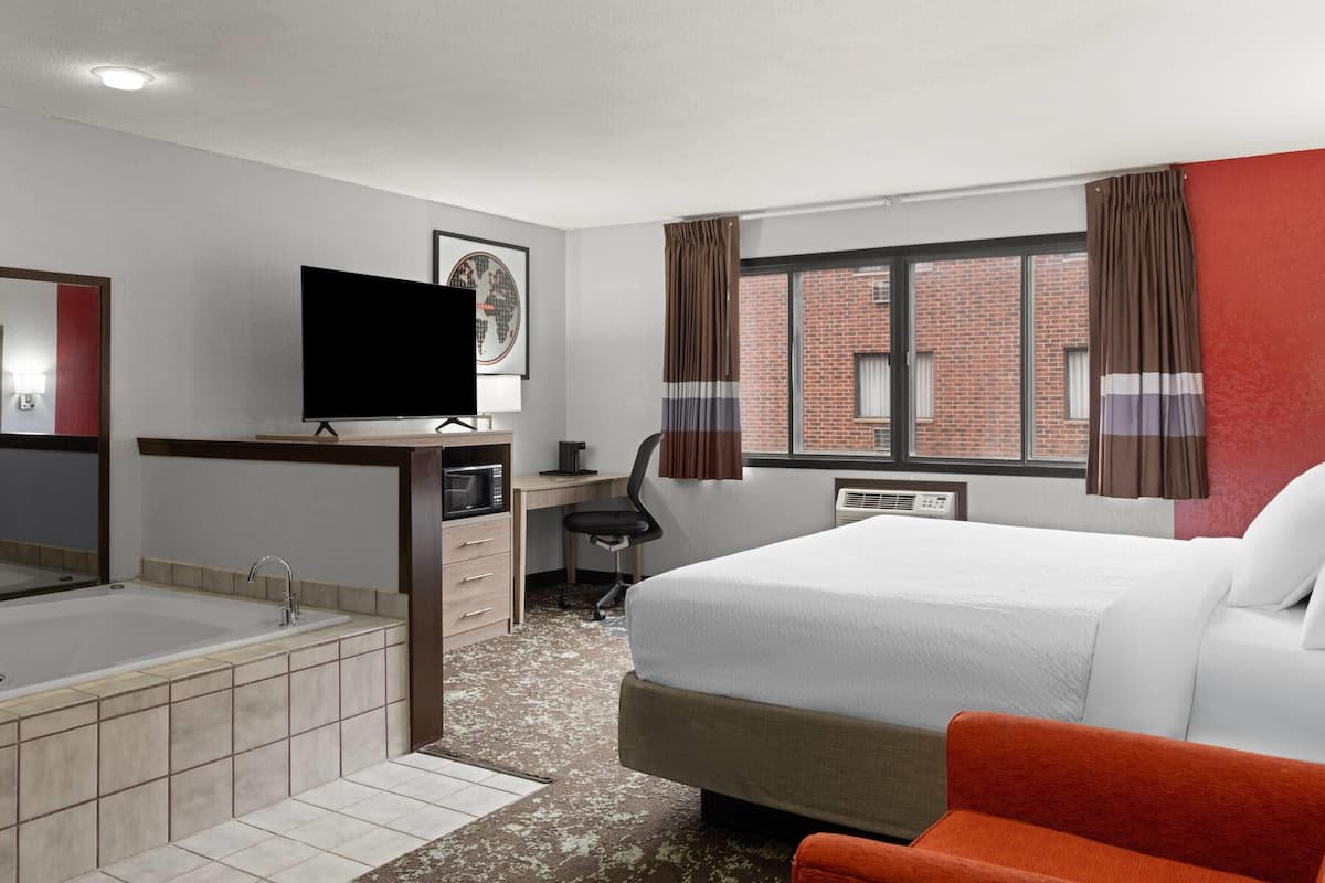 Una habitación de hotel con cama, escritorio con computadora, televisor, baño con bañera y ventana con cortinas.