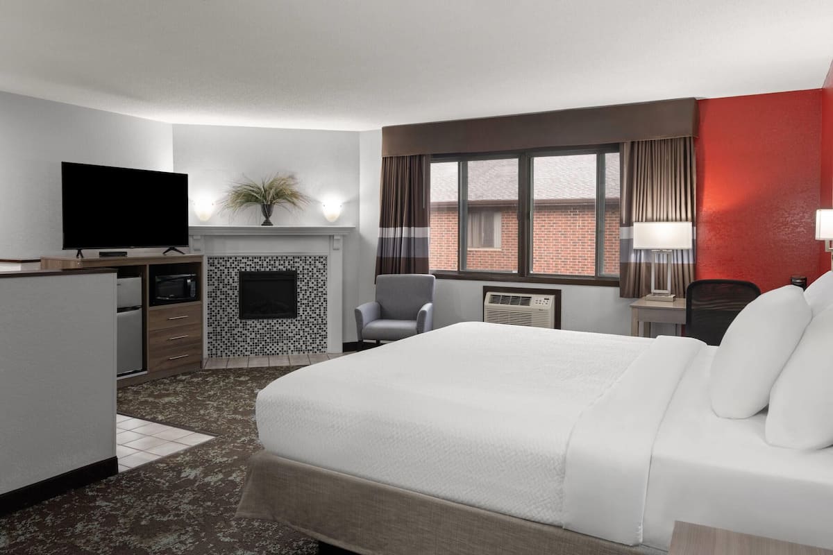 Una habitación de hotel con una cama, un televisor de pantalla plana, un escritorio pequeño con lámpara, una silla, una chimenea y una ventana con cortinas.