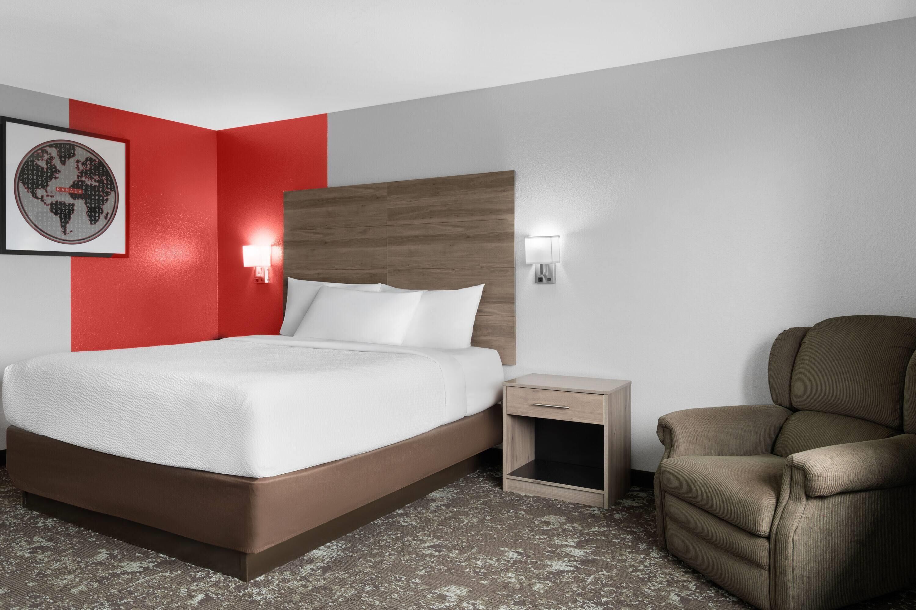 Quarto de hotel com uma cama, uma mesa de cabeceira, uma cadeira, uma luminária na parede e um quadro enquadrado.