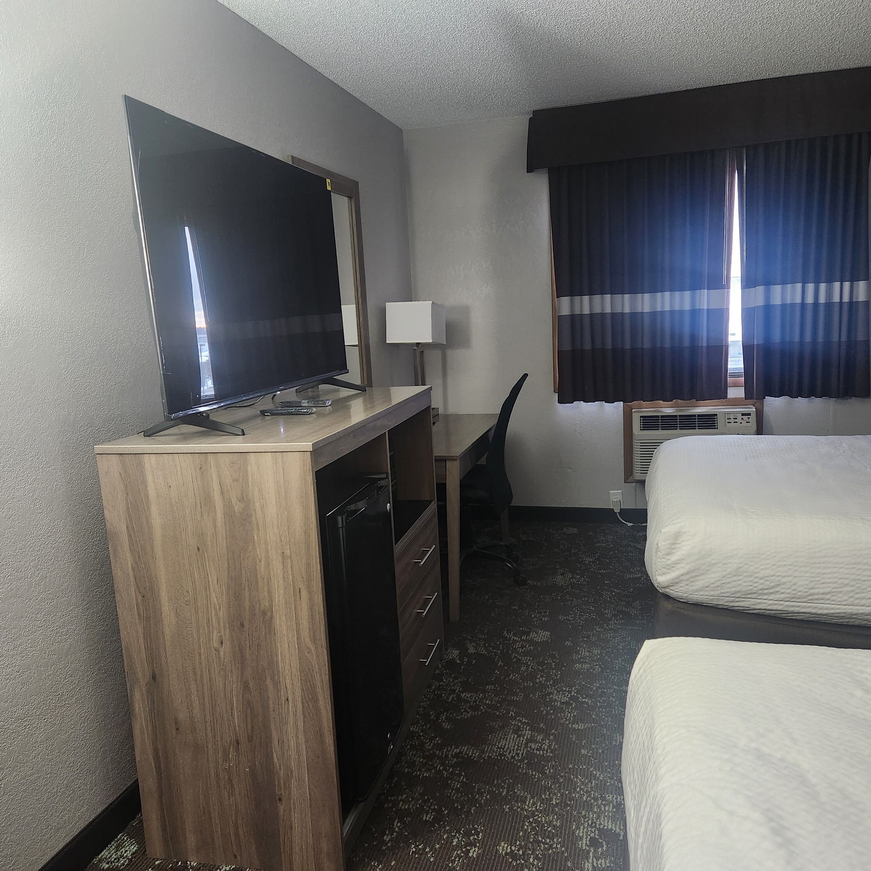 Ein Hotelzimmer mit einem Holztisch, einem Flachbildfernseher, einem Sessel, einem Bett und einem Fenster mit Vorhängen.