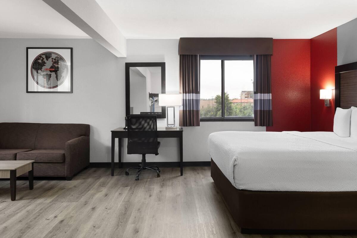 Una habitación de hotel con cama, escritorio, silla, mesita, espejo y ventana con cortinas.
