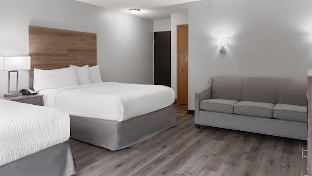 Habitación de hotel con cama doble, sofá gris, cabecero de madera, mesita de noche con lámpara y luz empotrada en la pared.