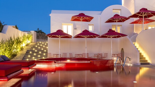 Un espace piscine agrémenté de parasols rouges, de chaises longues et d’un escalier menant à un bâtiment.