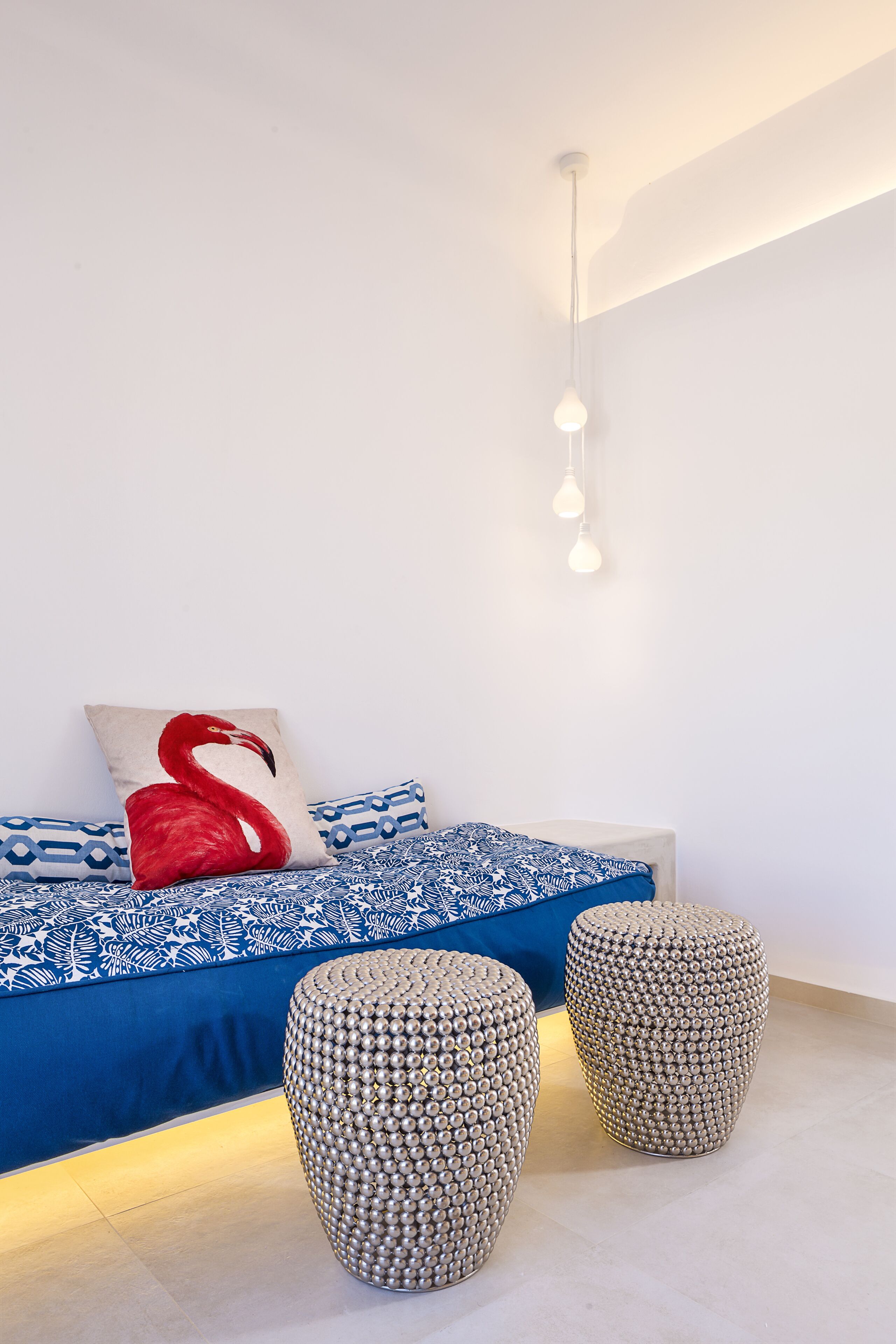 A modern bedroom with a blue patterned bedspread, a red and white swan pillow, and two metallic stools.