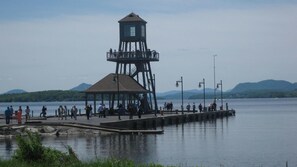 Un faro con una plataforma de observación y un muelle que se adentra en un lago tranquilo, rodeado de montañas.