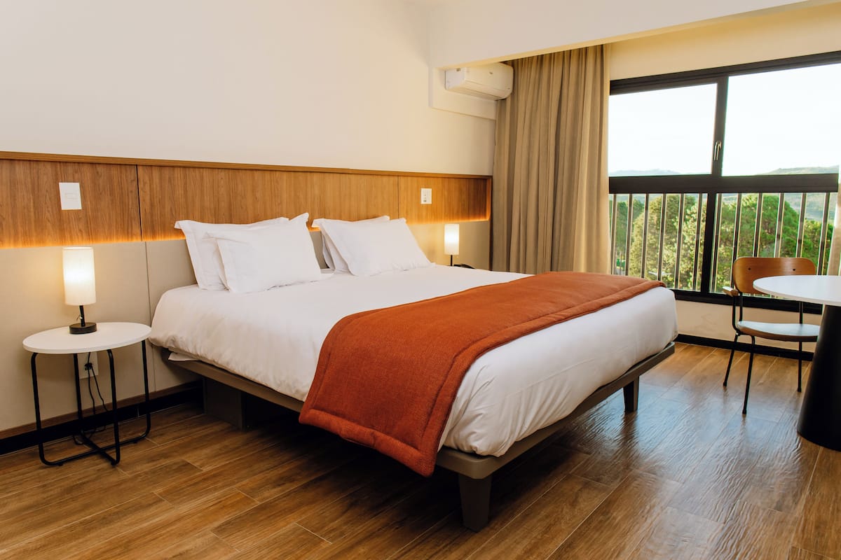 Ein Hotelzimmer mit einem Bett, zwei Nachttischen, einem Sessel, einem kleinen Tisch und einem Fenster mit Blick.