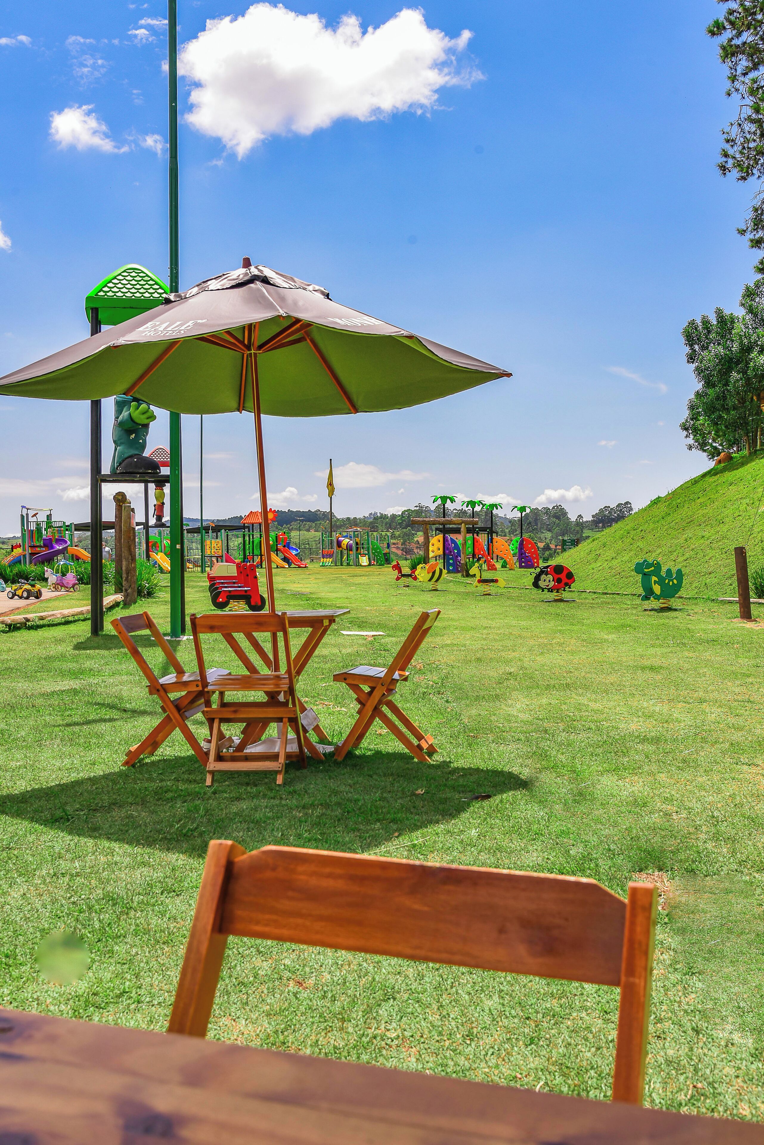 Un espacio al aire libre con césped, una mesa y sillas de madera bajo un paraguas verde, rodeado de juegos infantiles.