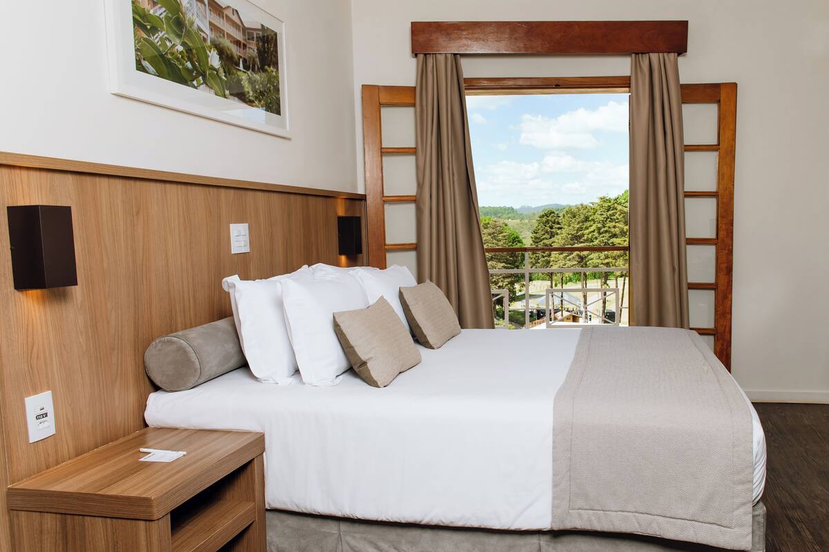 Ein Hotelzimmer mit Bett, Holz-Kopfteil, Nachttisch, Fenster mit Vorhängen und Blick ins Grüne.