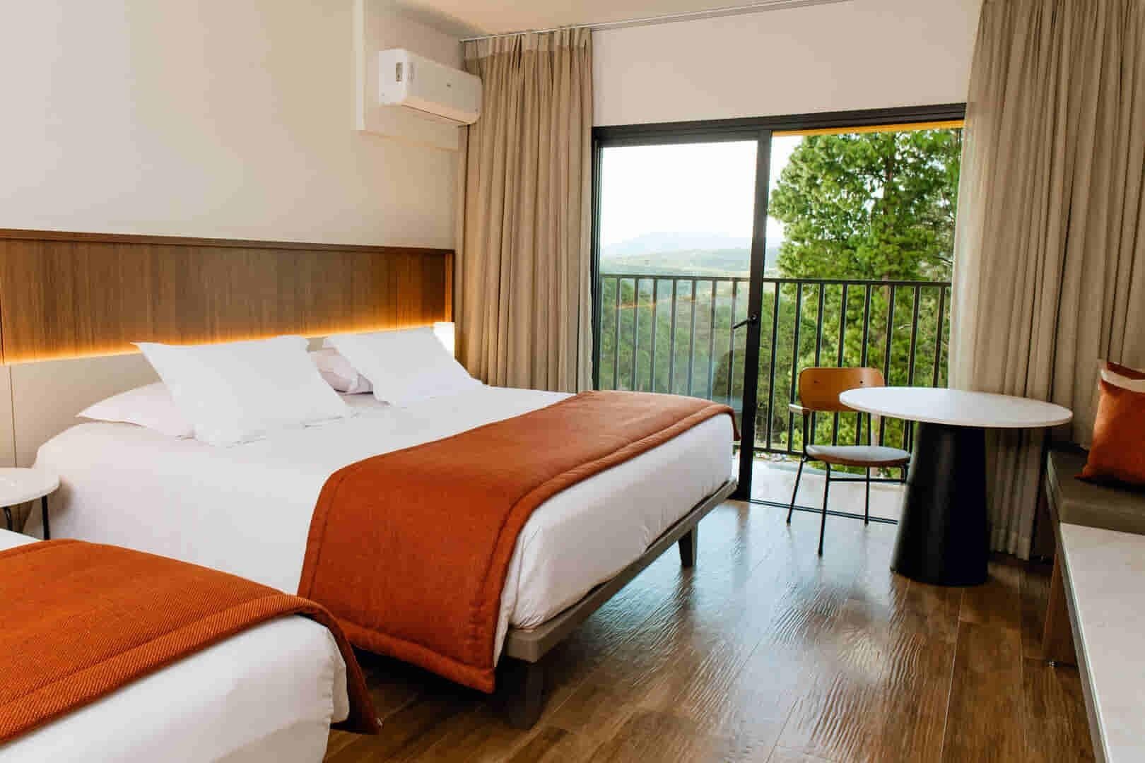 A hotel room with two beds, a balcony with a table and chairs, and a view of greenery.