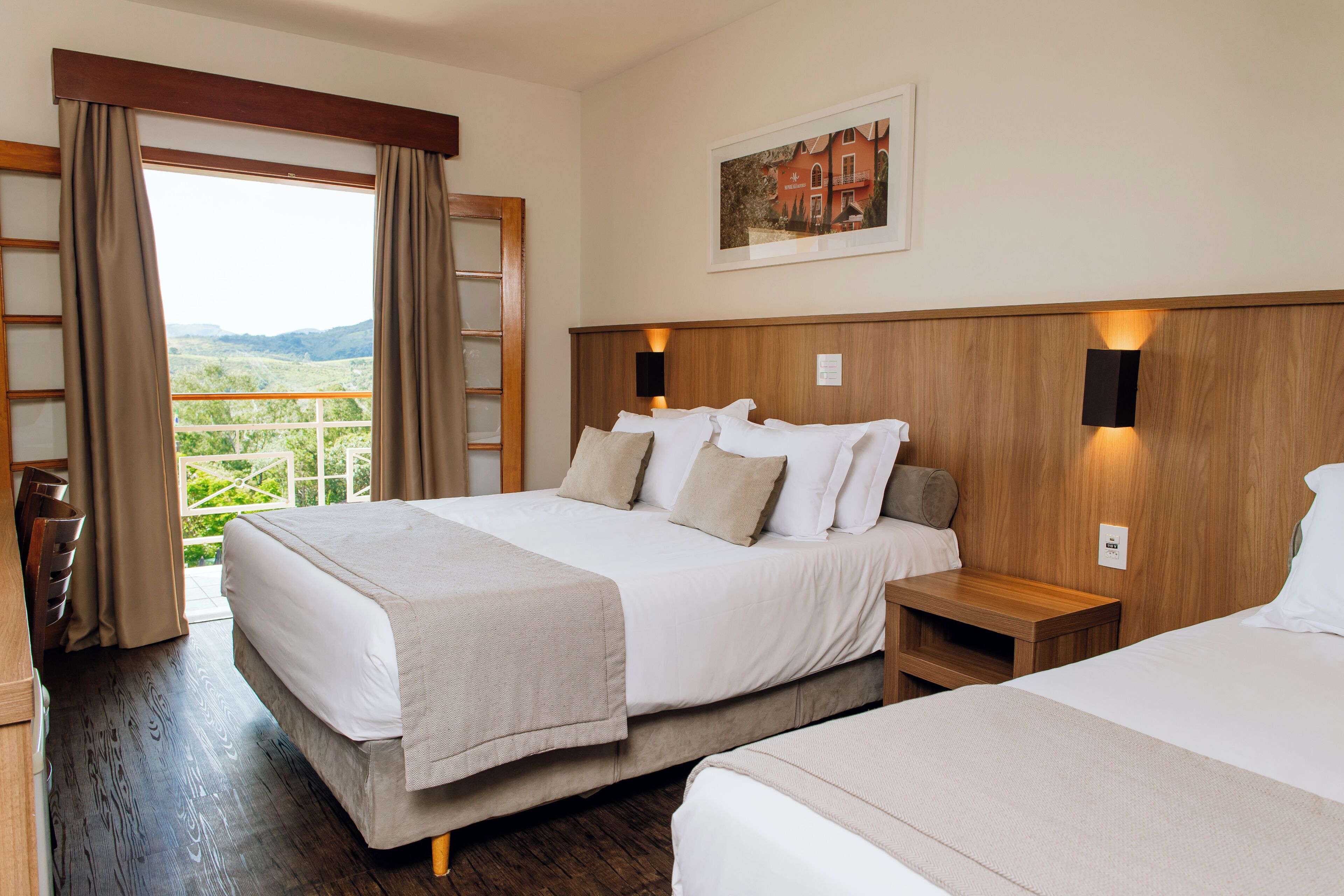 Habitación de hotel con dos camas, cabecero de madera, mesita de noche, un cuadro en la pared y vista a la vegetación por la ventana.