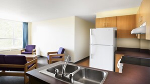 A compact kitchen with a sink, wooden cabinets, and a refrigerator. There are two purple chairs and a small table.
