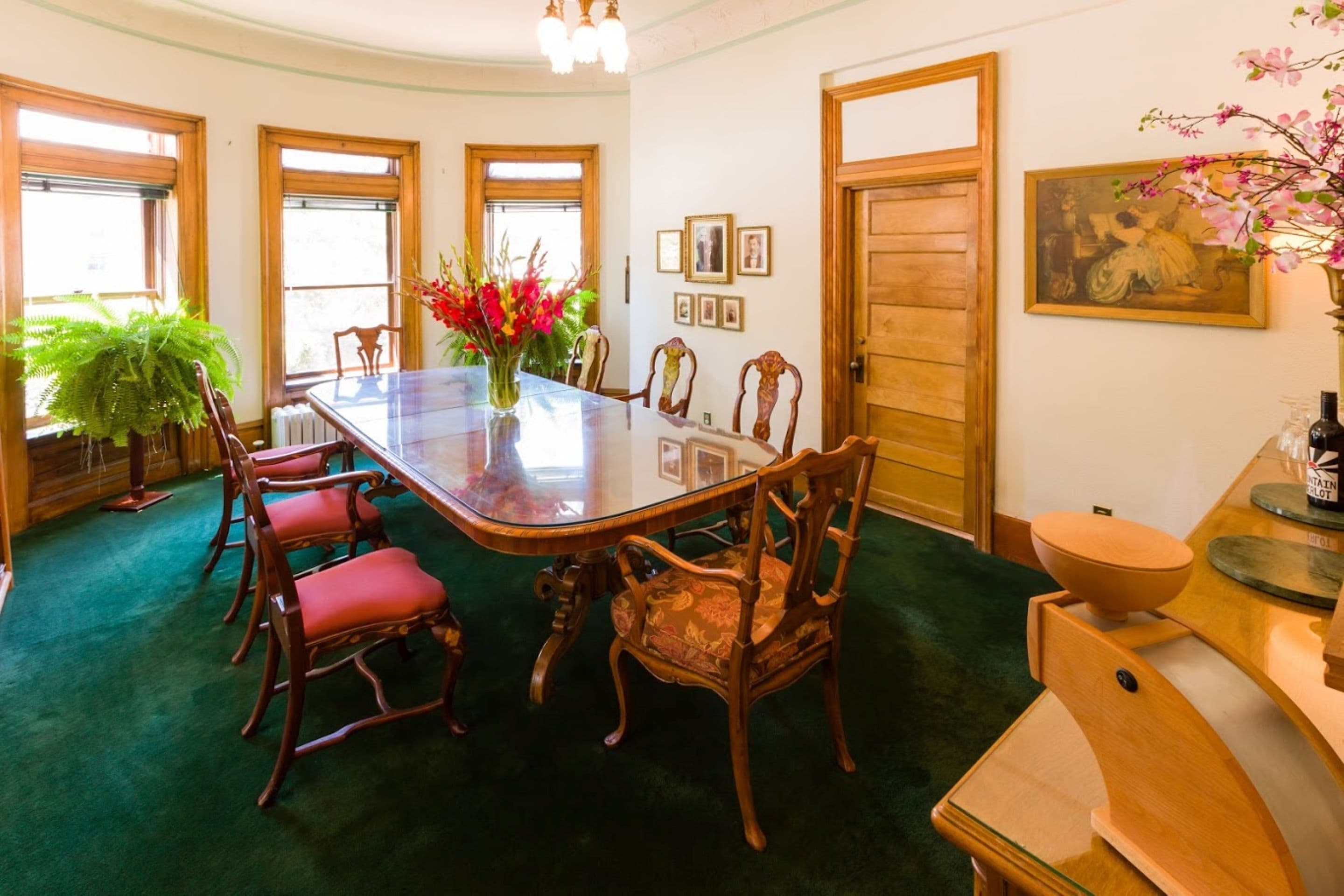 A dining area with a glass-top table surrounded by wooden chairs, a green carpet, and a large potted plant.