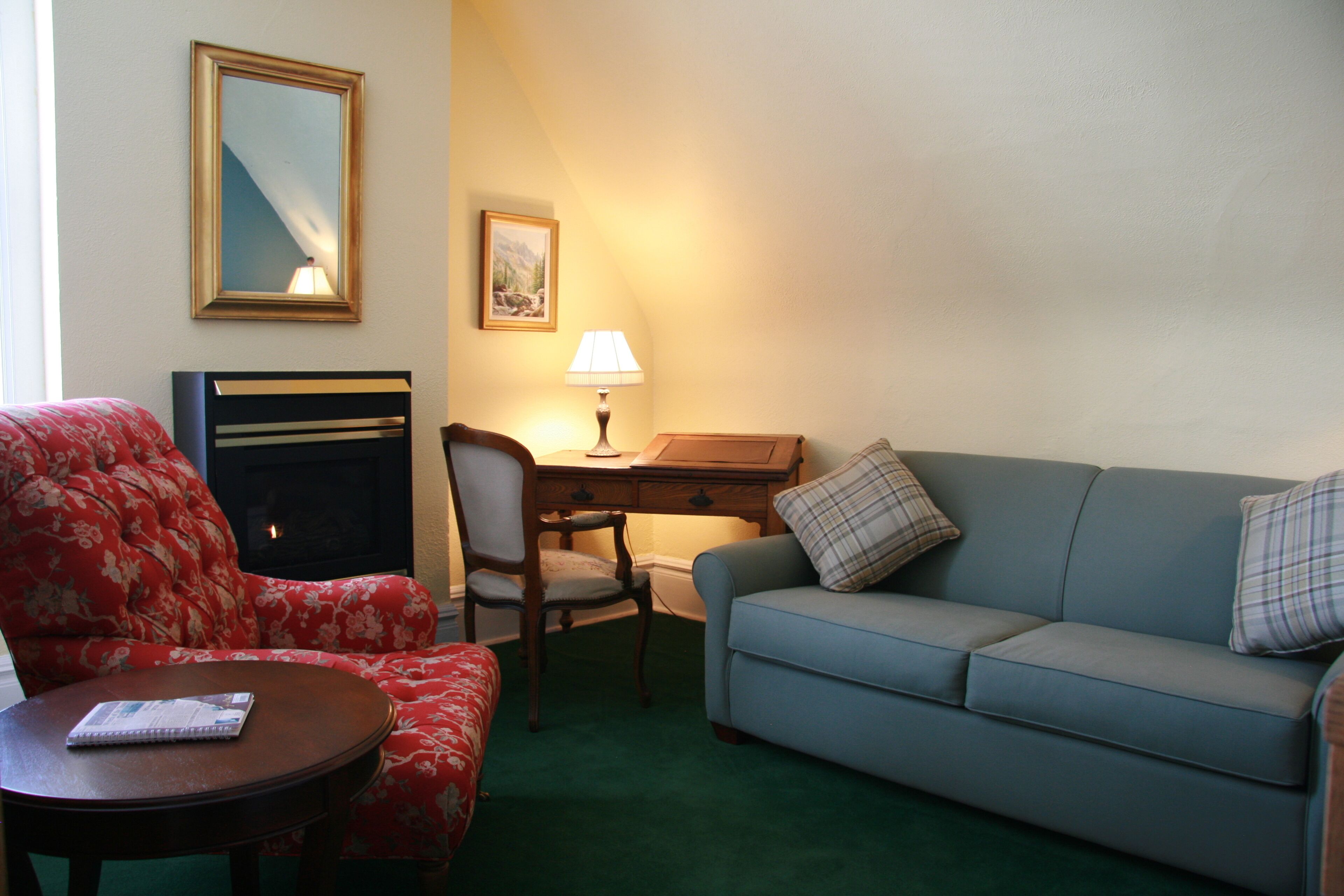 A room with a red floral armchair, a blue sofa with plaid cushions, a wooden desk, a lamp, a framed picture on the wall, and a fireplace.
