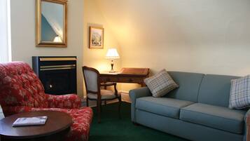 Une pièce comprenant un fauteuil à motifs floraux rouges, un canapé bleu avec des coussins à carreaux, un bureau en bois, une lampe, un tableau encadré accroché au mur et une cheminée.