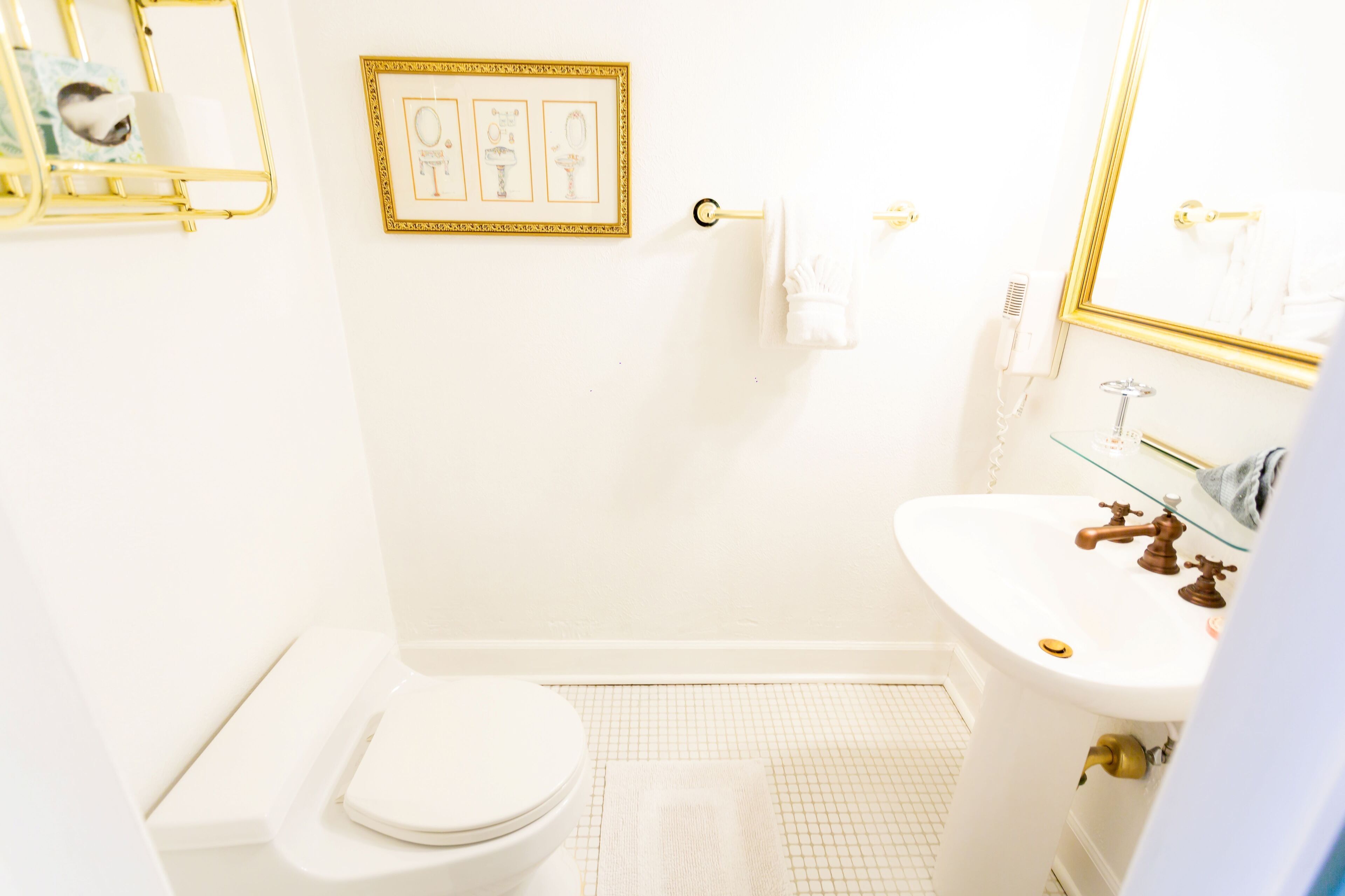 Un baño pequeño y blanco con inodoro, lavamanos y espejo. Hay un cuadro en la pared y un perchero para toallas.