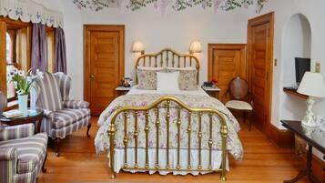A bedroom with a wooden bed, two bedside lamps, a floral bedspread, a striped armchair, and a small table with a vase of flowers.