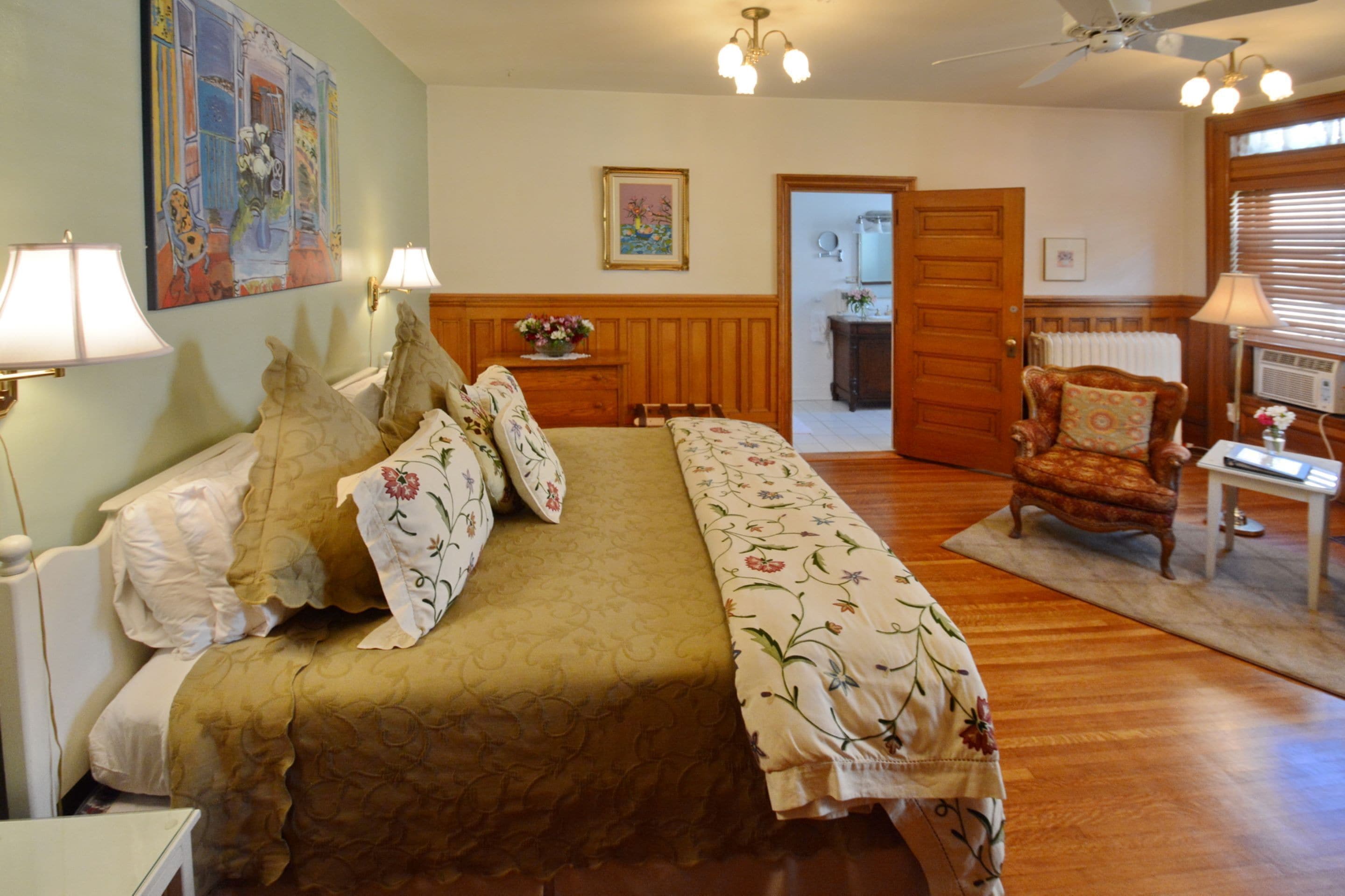 Quarto com uma cama grande, cabeceira de madeira, duas luminárias fixadas na parede, cômoda de madeira e uma cadeira.