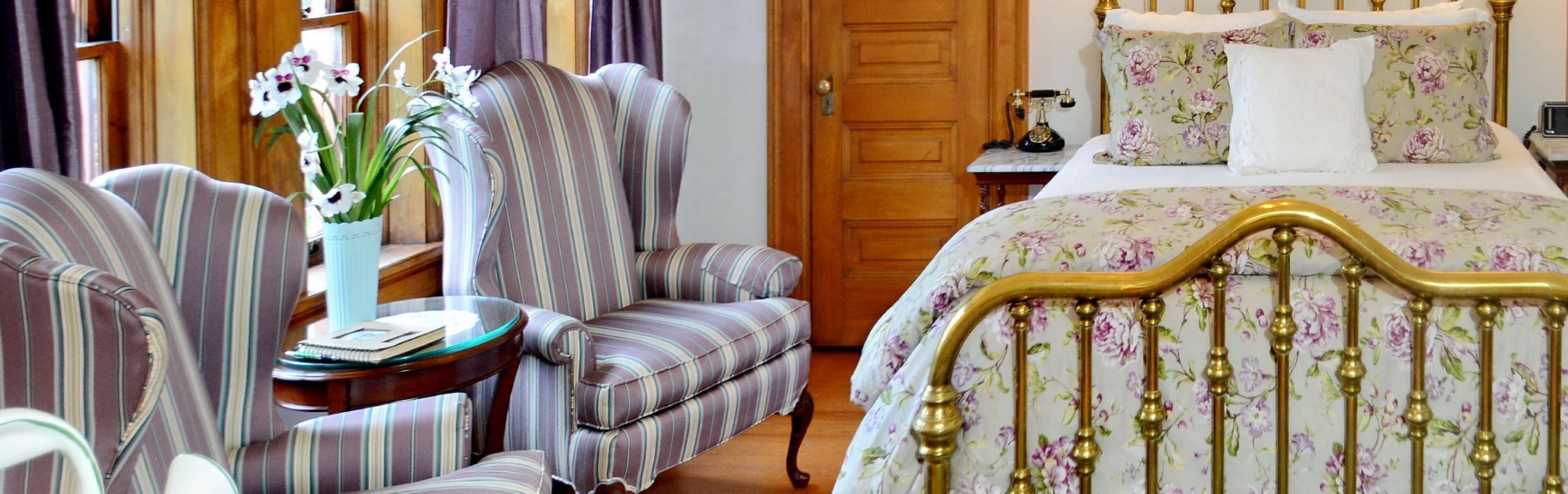 Quarto de hotel com uma cama, duas poltronas listradas, uma mesinha com um vaso de flores e uma porta de madeira.