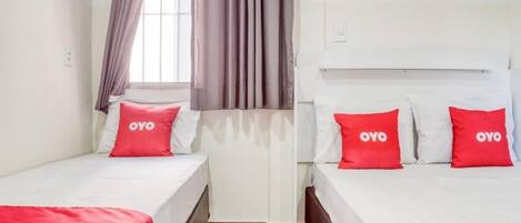 Habitación de hotel con dos camas individuales, cada una con una almohada roja con la marca OYO, ropa de cama blanca y una manta roja. Hay una ventana con cortinas, un radiador y suelo de baldosas.