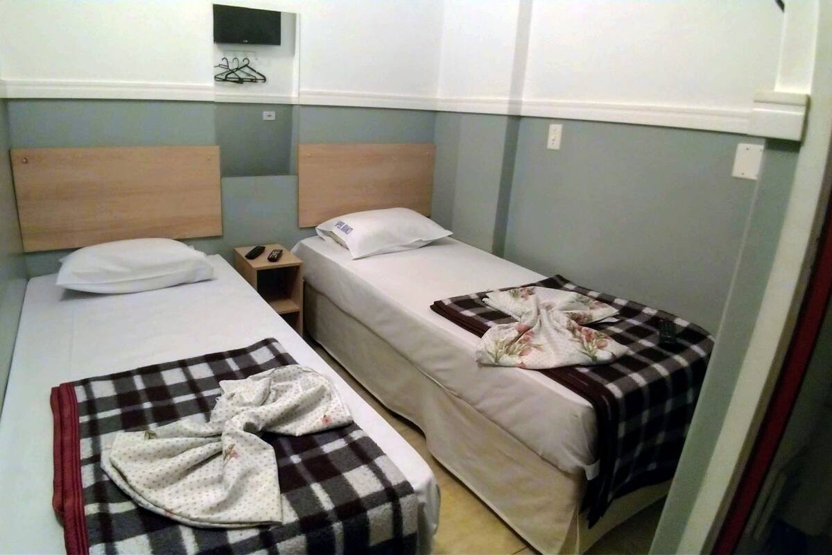 Ein kleines Zimmer mit zwei Einzelbetten, jeweils mit Kopfkissen und Decke. Ein kleiner Nachttisch bietet Platz für ein Telefon und eine Fernbedienung. Das Zimmer ist schlicht und zweckmäßig eingerichtet, mit hellen Wänden und einem Holz-Kopfteil.
