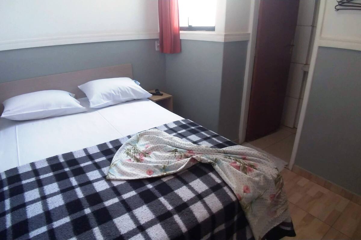 Ein ordentlich bezogenes Bett mit kariertem Überwurf und floralem Bettbezug, ein Nachttisch mit Lampe und ein Fenster mit Vorhängen.