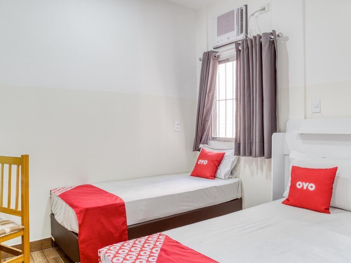 A small, clean room with two beds, a wooden chair, and a window with curtains.