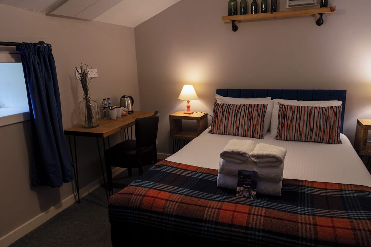 A hotel room with a bed, a desk, a chair, a lamp, and a shelf with bottles.