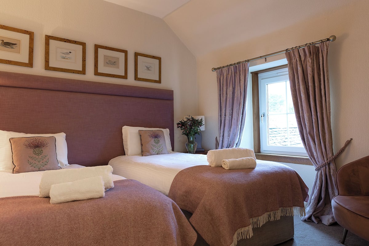 A hotel room with two beds, a large window with curtains, a chair, and framed pictures on the wall.