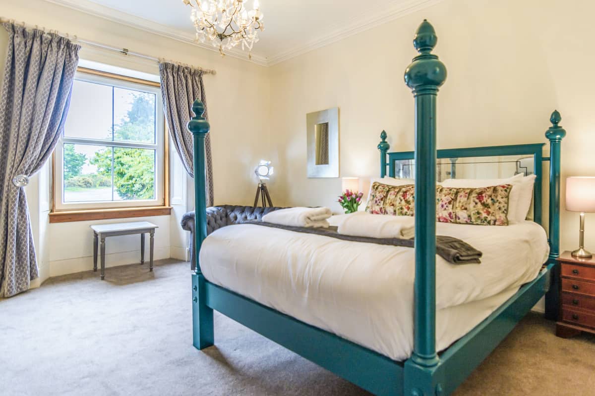 A four-poster bed with a floral bedspread, a bedside table, a lamp, and a window with curtains.