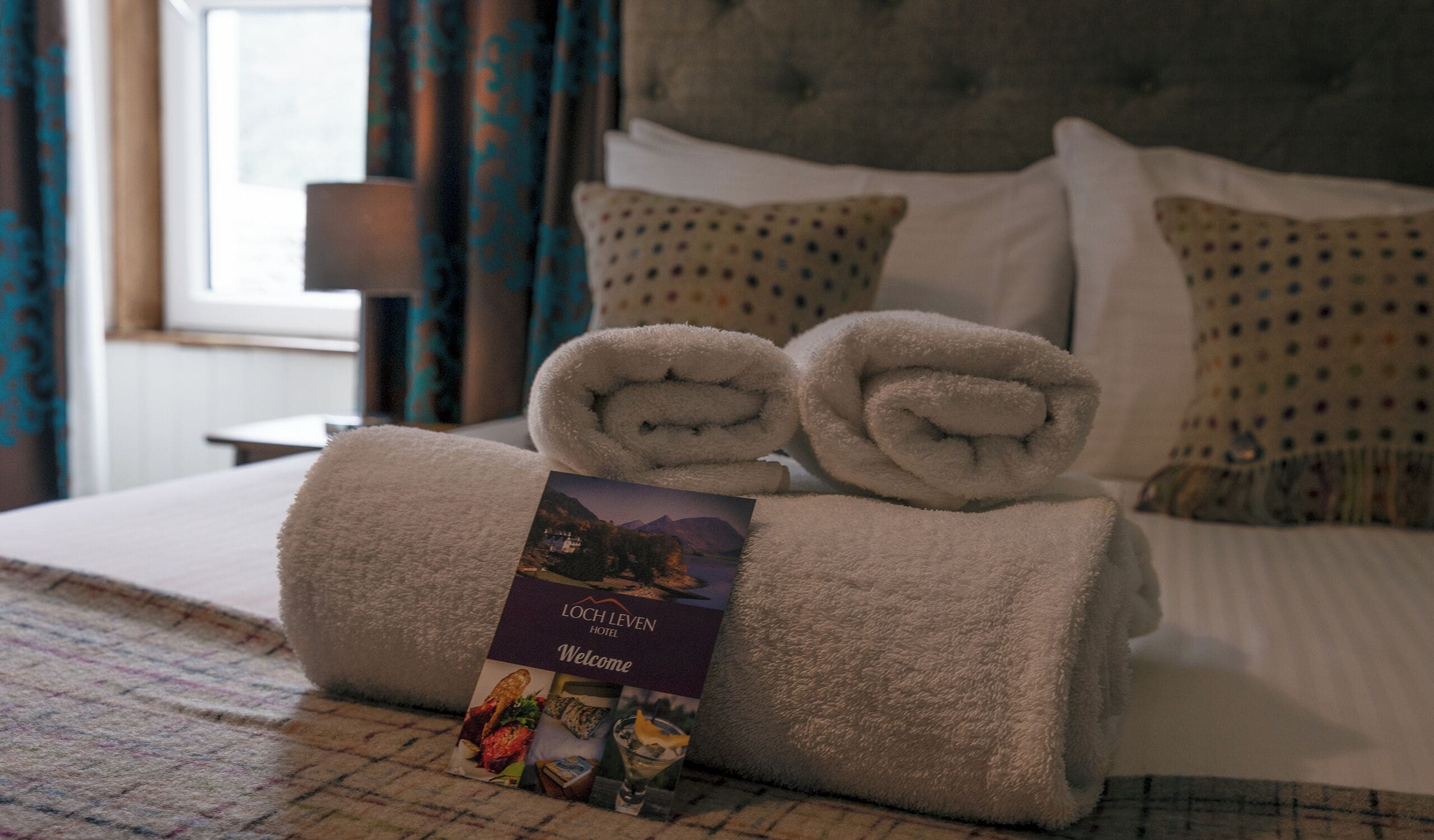 A neatly made bed with rolled towels and a welcome brochure.