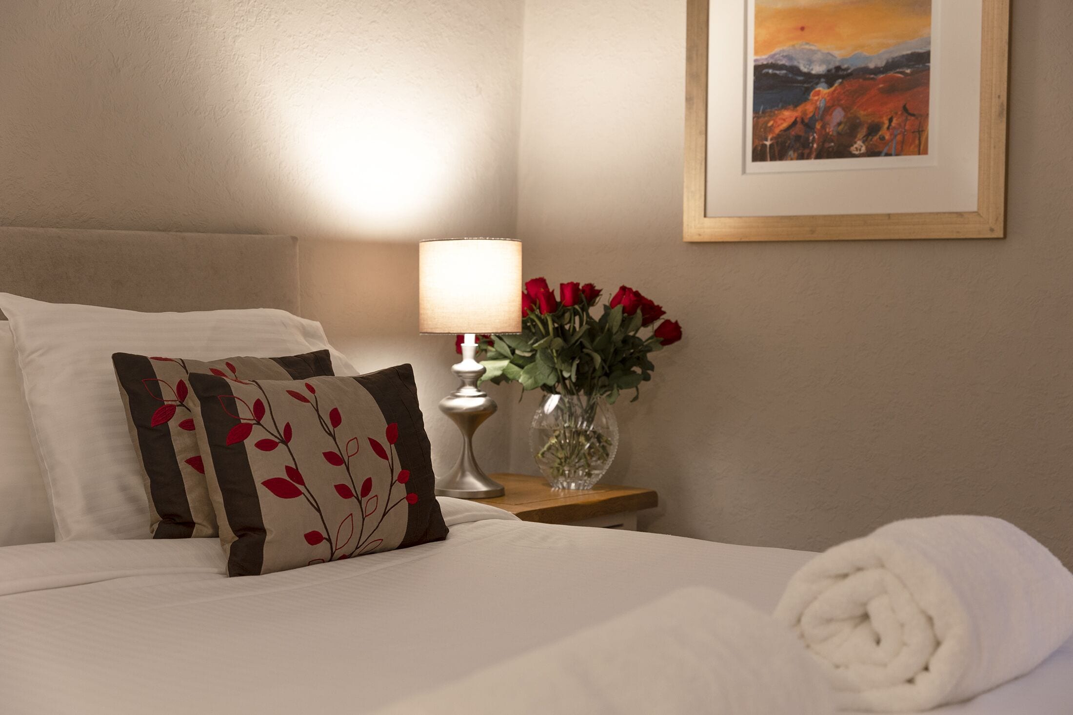 A neatly made bed with white linens, a vase with red roses, and a framed landscape painting on the wall.