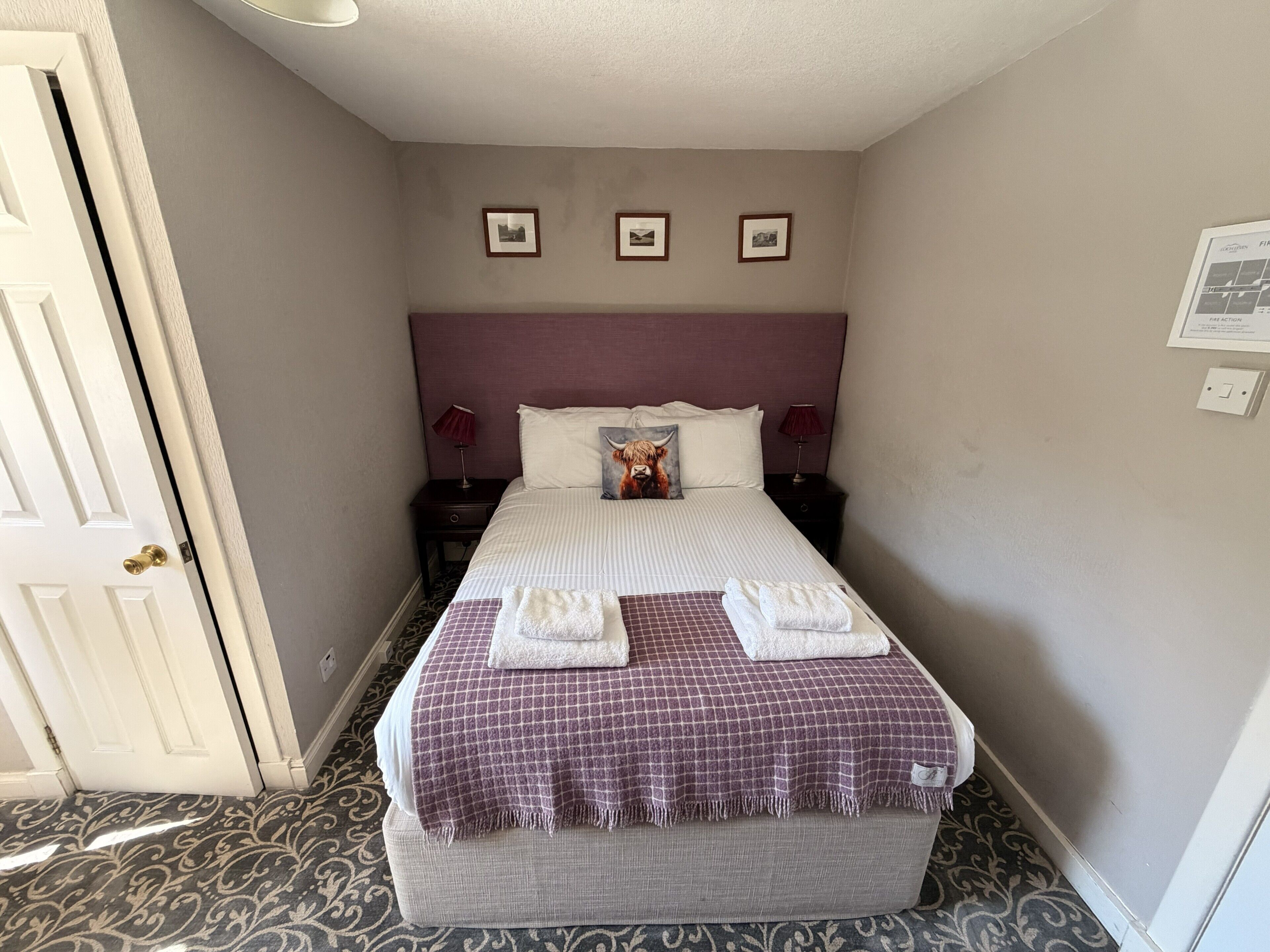 Une chambre d’hôtel avec un lit, une tête de lit, une table de chevet, une lampe, un tableau accroché au mur et une porte.