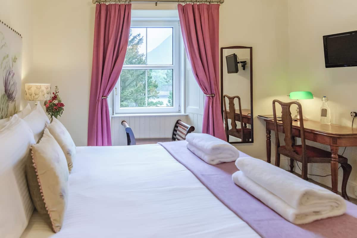 A neatly made bed with white linens, a wooden chair, a desk with a lamp, and a window with pink curtains.