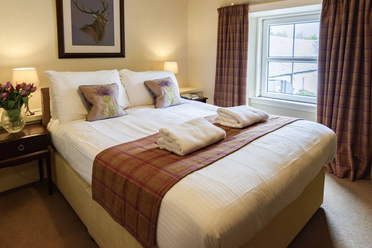 A neatly made bed with white linens, a plaid blanket, and two pillows with floral designs. There is a framed deer head on the wall, a bedside table with a lamp and a vase of flowers, and a window with curtains.