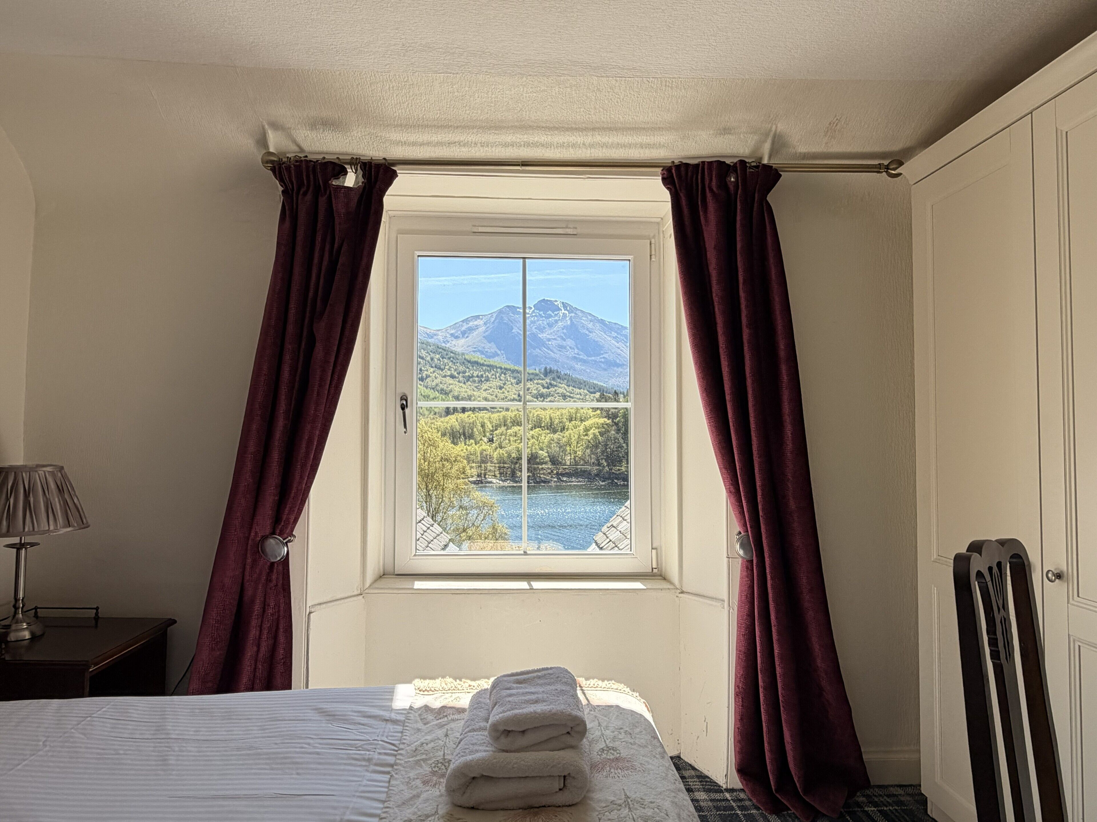 A bedroom with a bed, a window with curtains, a lamp, and a view of a mountain and lake.