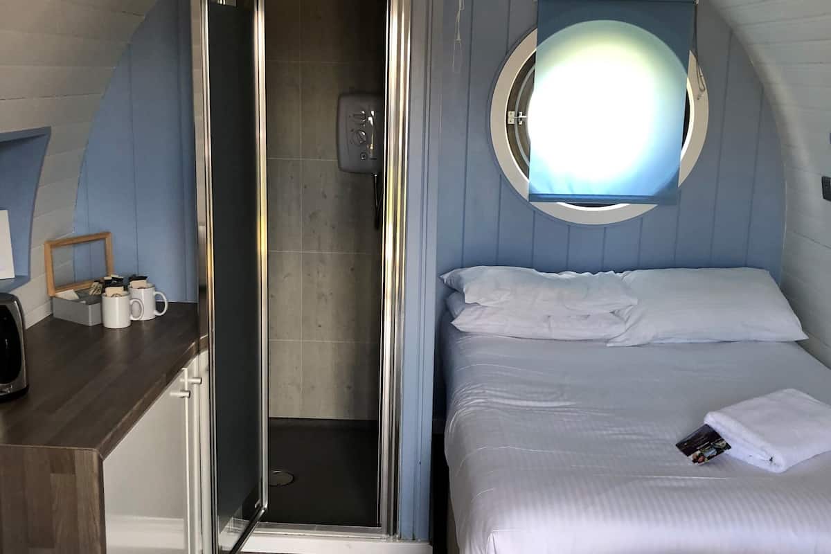 A compact room with a bed, a small desk, a shower, and a round window.