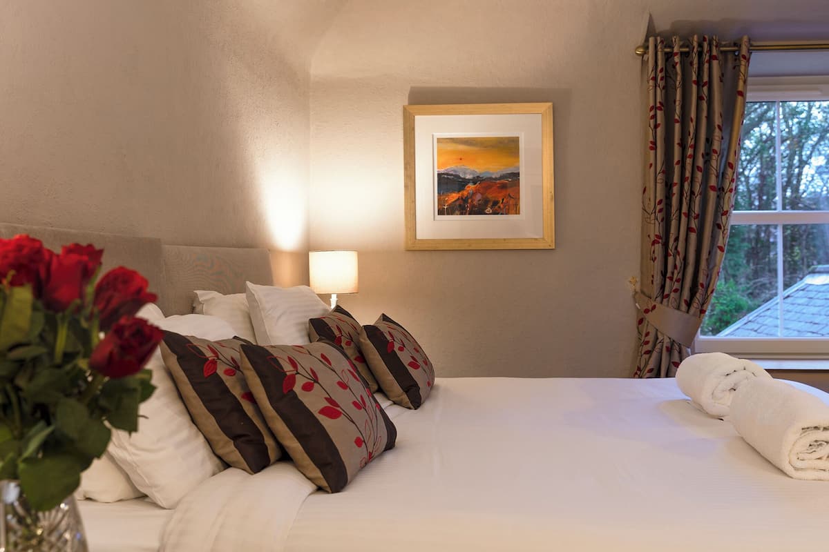 A neatly made bed with white linens, a vase of red roses, and a framed landscape painting on the wall.