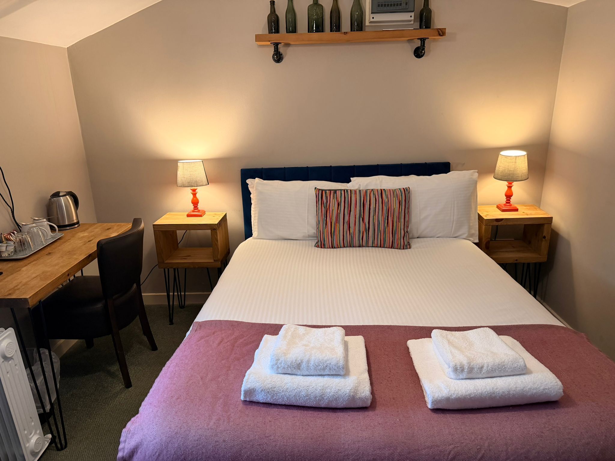 Habitación de hotel con cama, dos mesitas de noche con lámparas, un escritorio con hervidor y una silla.