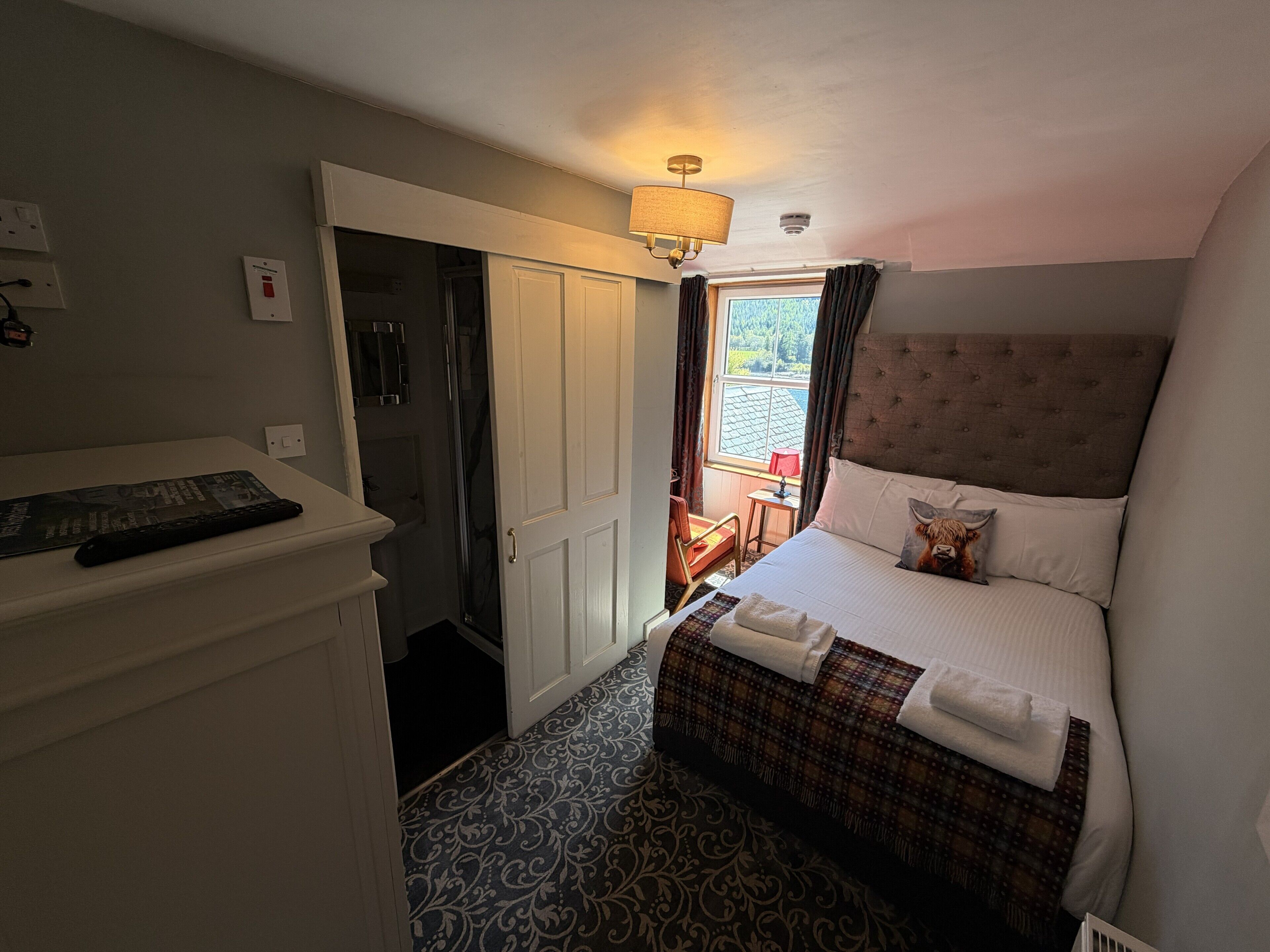 Habitación de hotel con cama, mesita de noche, lámpara, zona de ducha y vistas al exterior.