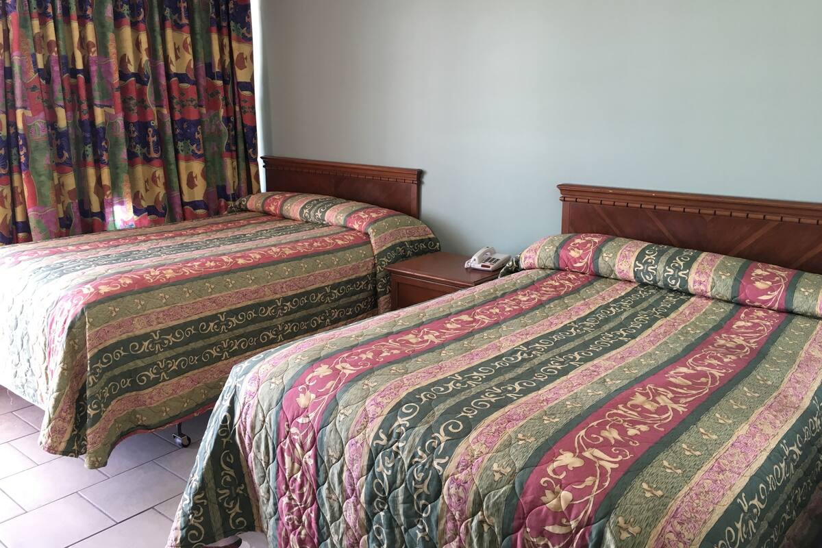 A hotel room with two beds, a wooden headboard, a bedside table with a phone, and a curtain with a colorful pattern.