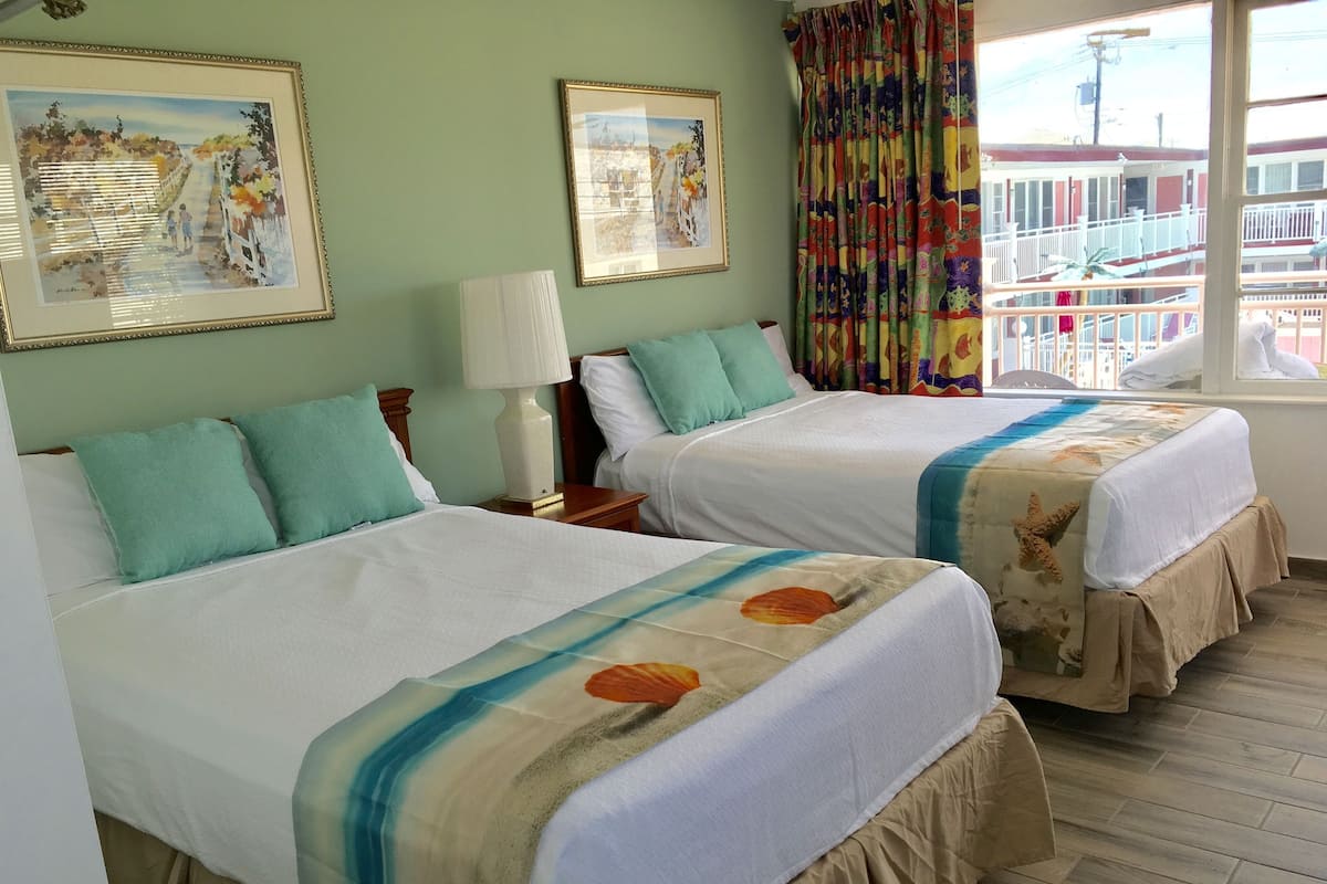 A hotel room with two beds, each with a white bedspread and teal pillows. There are framed pictures on the wall, a window with curtains, and a view of a balcony outside.
