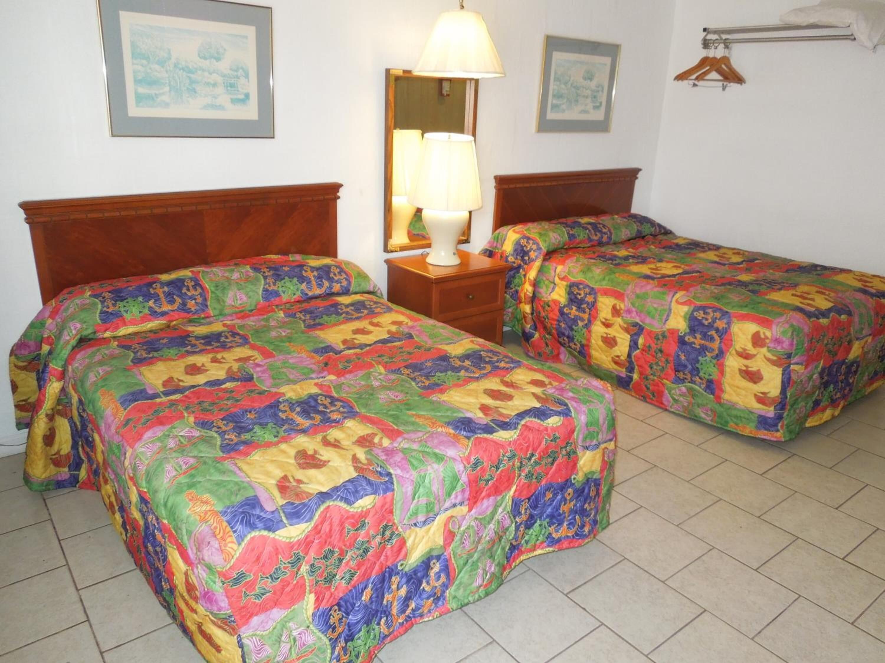 Habitación de hotel con dos camas, cada una con un edredón colorido, una mesita de noche con lámpara y un perchero.