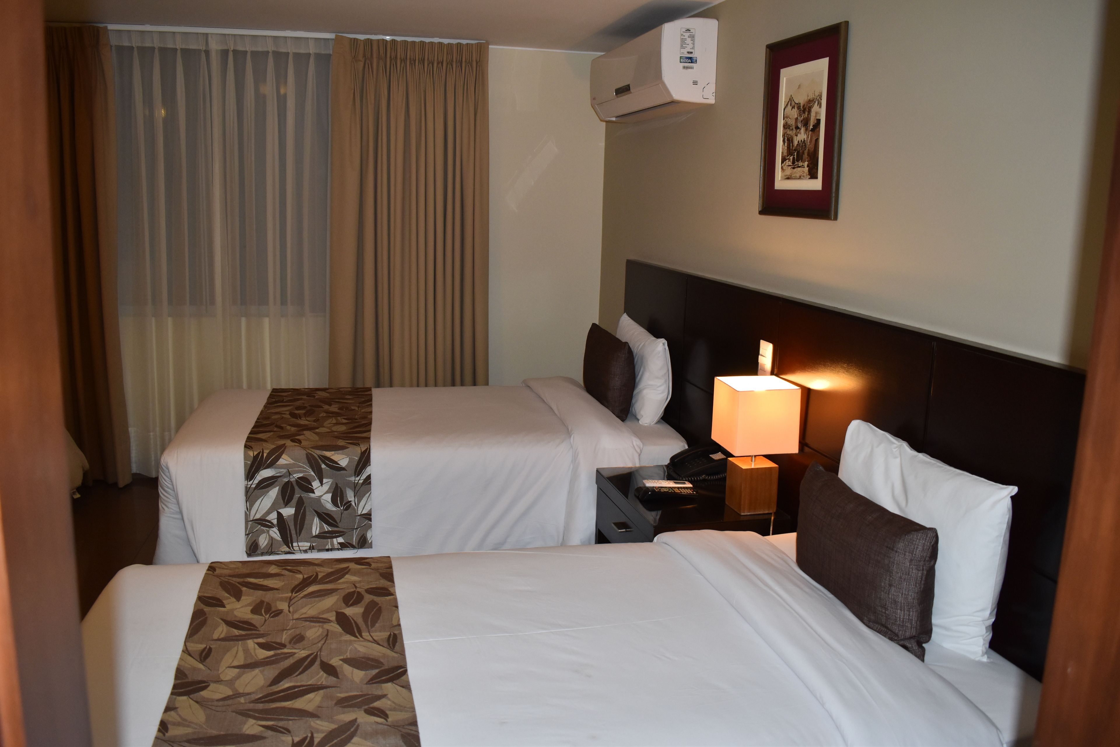 A hotel room with two beds, a nightstand with a lamp, an air conditioner, and a framed picture on the wall.