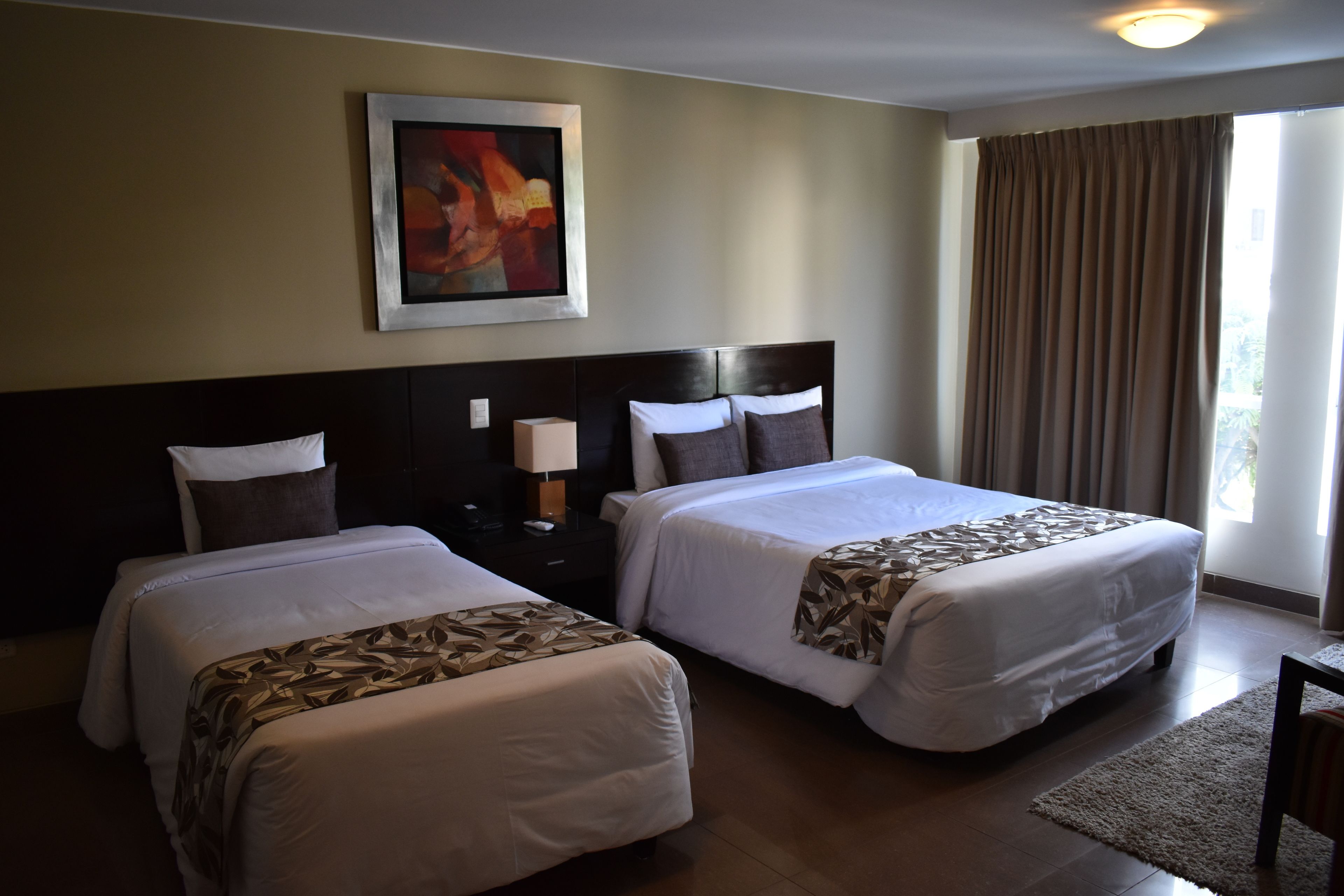 Habitación de hotel con dos camas, una mesita de noche con lámpara, un cuadro en la pared y una ventana con cortinas.