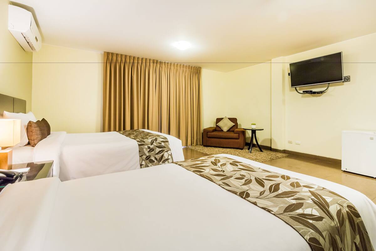 A hotel room with two beds, a TV, a desk with a lamp, a chair, and a small table.