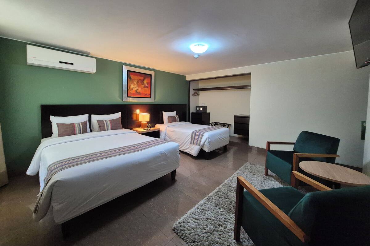 A hotel room with two beds, a desk, a chair, a small table, a lamp, and a painting on the wall.