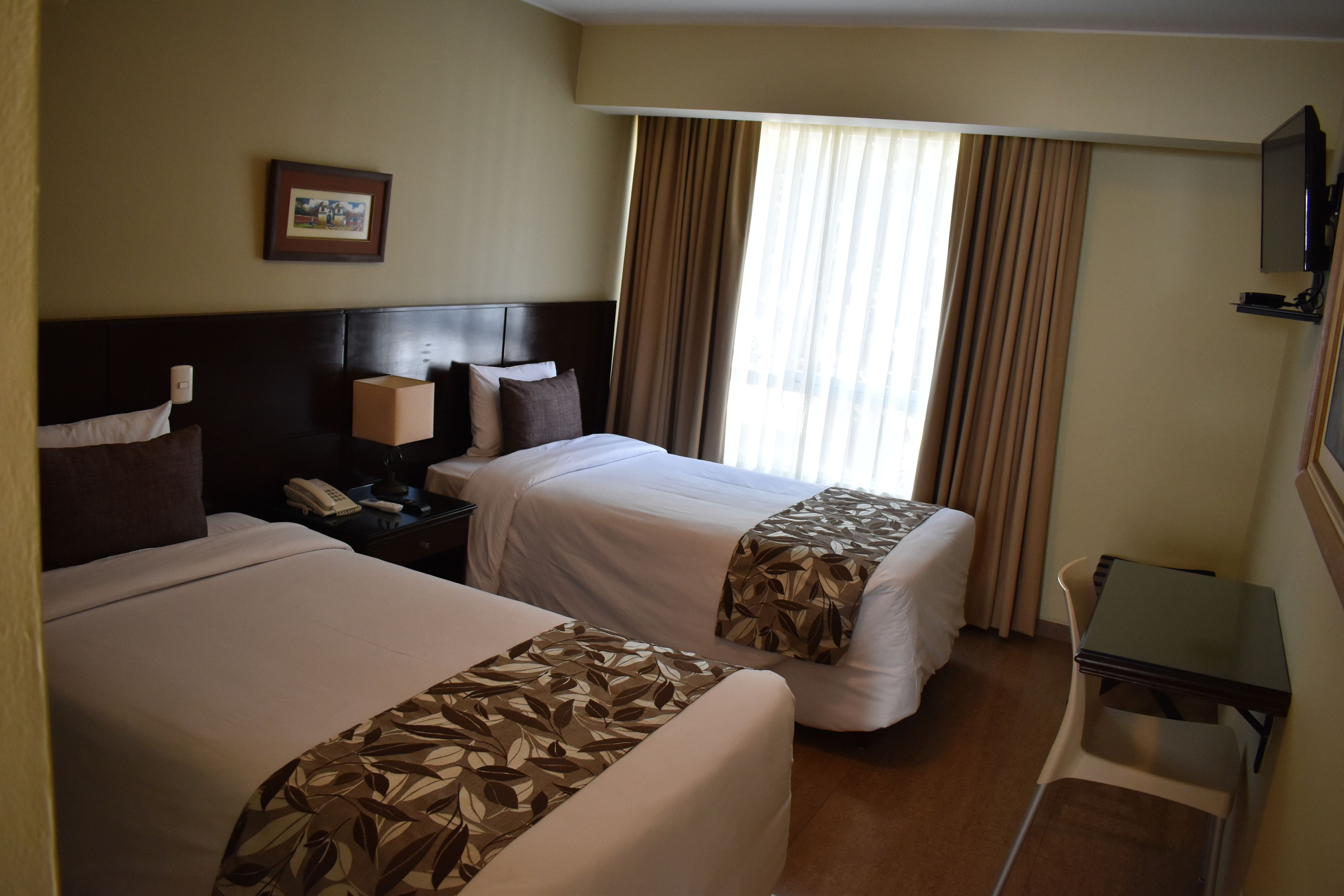 A hotel room with two beds, a bedside table with a lamp, a small desk, and a TV mounted on the wall.