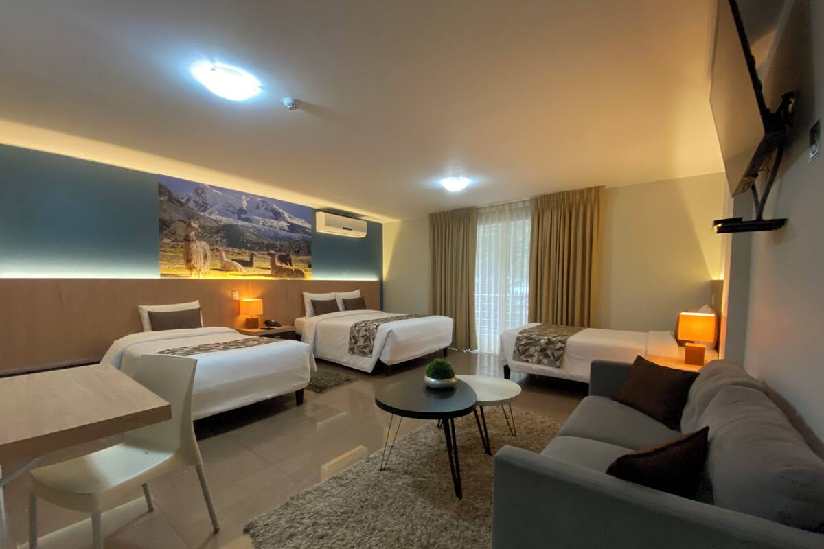 A hotel room with two single beds, a sofa, a small table, and a chair. There is a painting on the wall and an air conditioning unit above the beds.