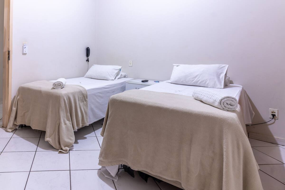 A hotel room with two single beds, each with a white bedspread and pillow, a bedside table with a phone, and a power outlet on the wall.