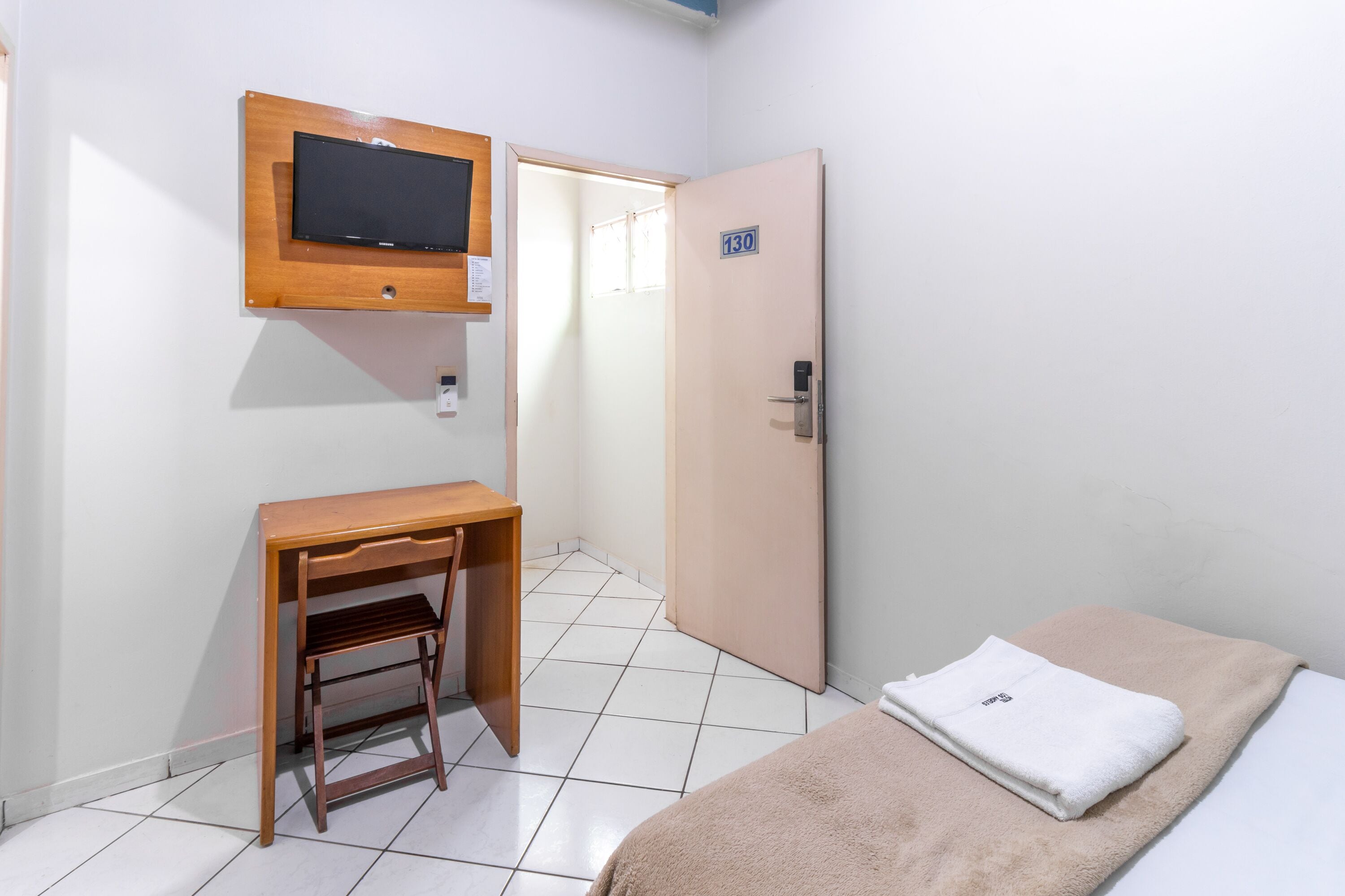A hotel room with a bed, a desk, a chair, a television mounted on the wall, and a door with a number sign.
